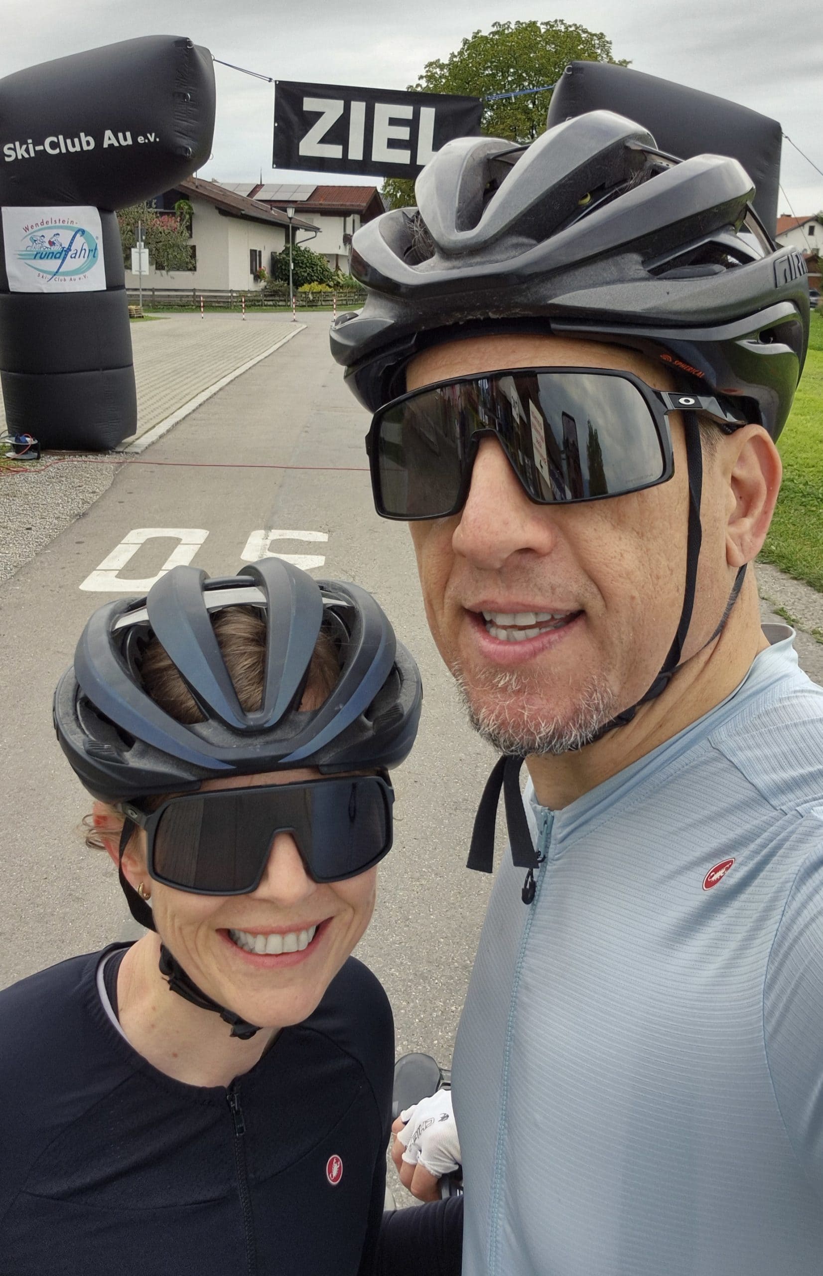 Zwei Personen in Fahrradkleidung machen vor dem Zielbogen einer Radsportveranstaltung ein Selfie